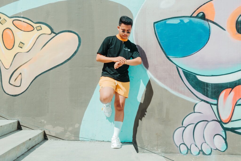 Full body of serious Asian male in stylish clothes and sunglasses looking at time on wristwatch while leaning on graffiti wall